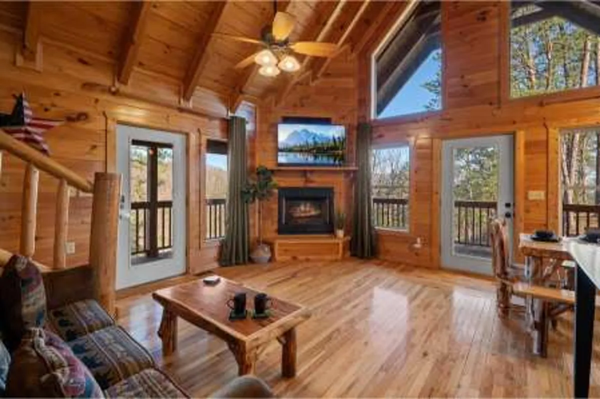 Inside the cabin, wooden beams frame a warm, welcoming living area with a fireplace, large windows, and a coffee table. Ample natural light pours in, and the windows offer views of the outdoors, reinforcing a connection to the mountain landscape.