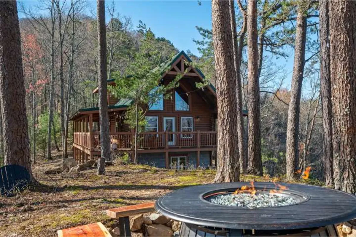 An inviting exterior view of the log cabin with a fire pit surrounded by trees, ideal for outdoor gatherings and evenings spent storytelling, with a sense of seclusion and connection to nature.