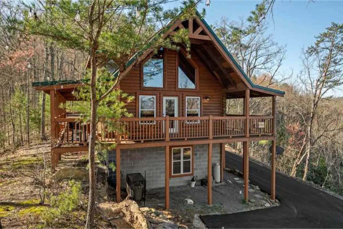 The image features a charming cabin nestled amid lush trees, showcasing a spacious wooden balcony, large windows that invite natural light, and a sturdy stone foundation. The tranquil setting highlights the inviting outdoor spaces perfect for relaxing or gathering, with hints of mountain vistas visible from the balcony.