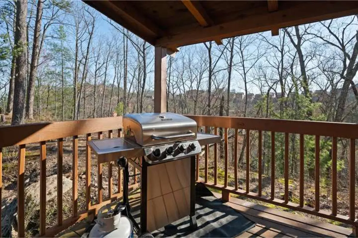 An exterior view focusing on a wooden deck with a grill, surrounded by trees and a wooden railing. The scene suggests a relaxed evening atmosphere ideal for outdoor dining, barbecues, and socializing with friends while enjoying the natural surroundings.