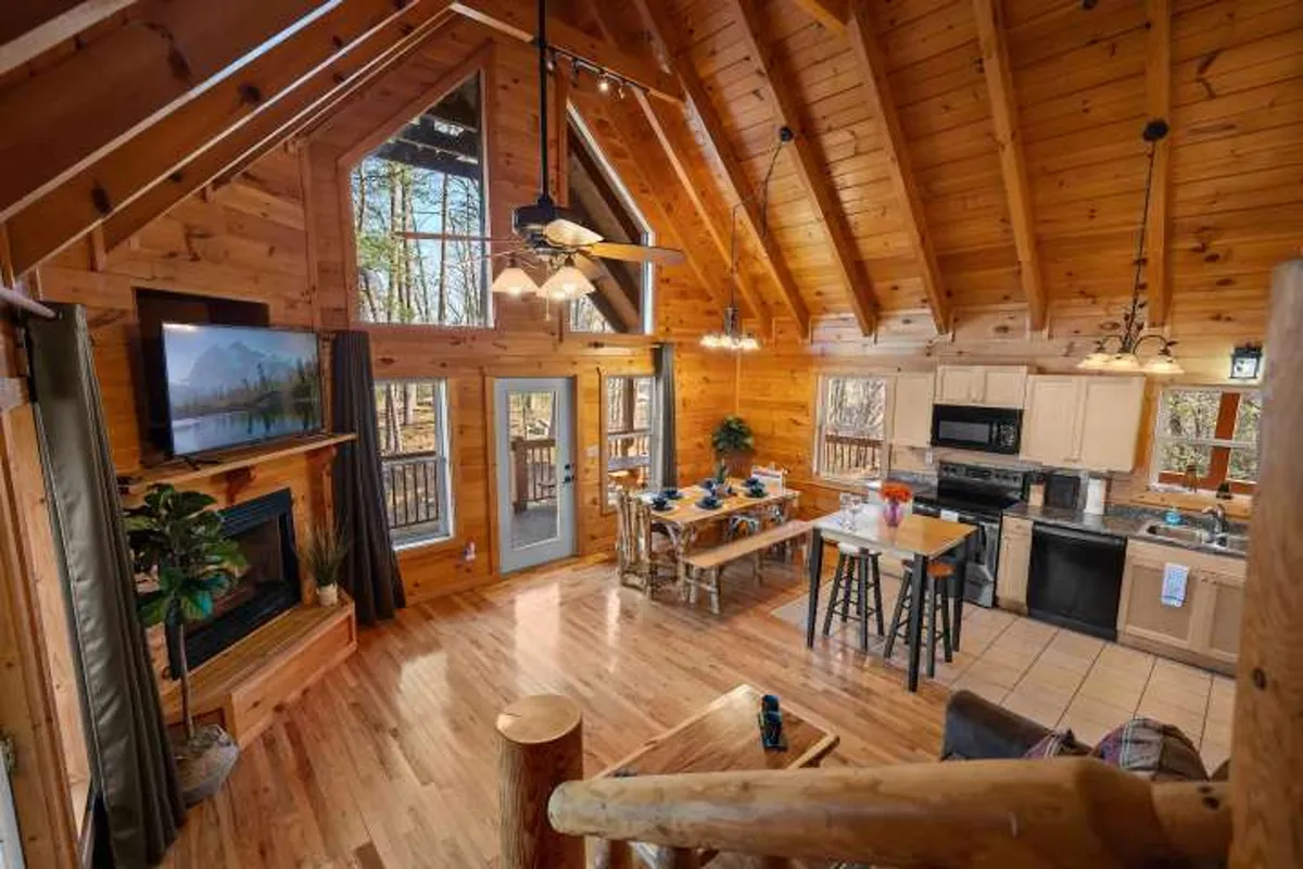 A wide shot of the living and dining area with large windows, exposed wooden beams, a welcoming fireplace topped with a TV, and a dining table set for a group meal. The space exudes a sense of warmth, making it ideal for family gatherings, game nights, or simply relaxing together.