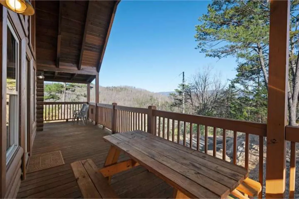A wooden picnic table sits on a deck with wooden railings, overlooking a sweeping mountain view beneath a clear blue sky. The setting is ideal for outdoor dining, relaxing, and enjoying the peacefulness of the mountains.