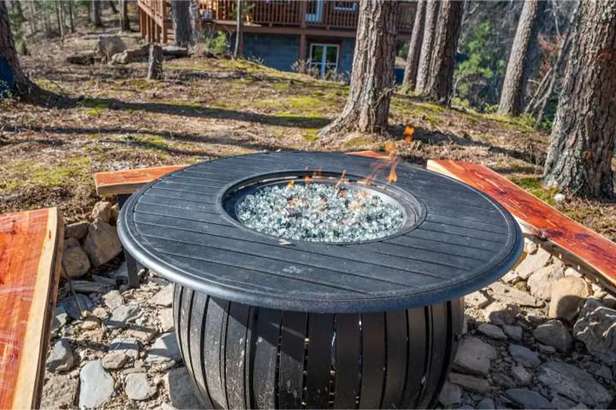 A close-up view of a modern fire pit encircled by seating, set on a patio with a backdrop of trees. The ambiance is warm and inviting, suggesting intimate conversations and laughter as evening falls.