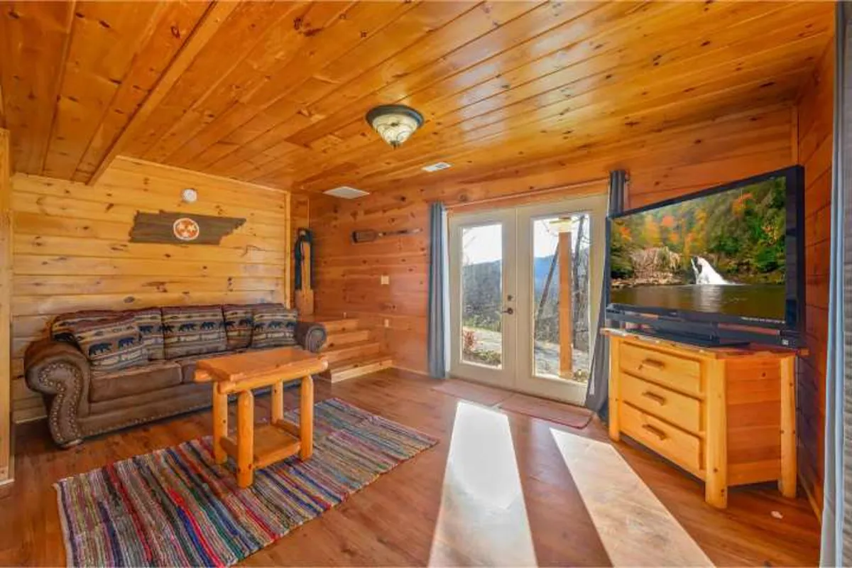 A cozy interior scene with a sofa, coffee table, and television set beside double doors that likely lead out to a porch or deck. The arrangement suggests a seamless transition from indoor comfort to outdoor relaxation, and hints at activities like reading by the fire or stargazing from the porch.