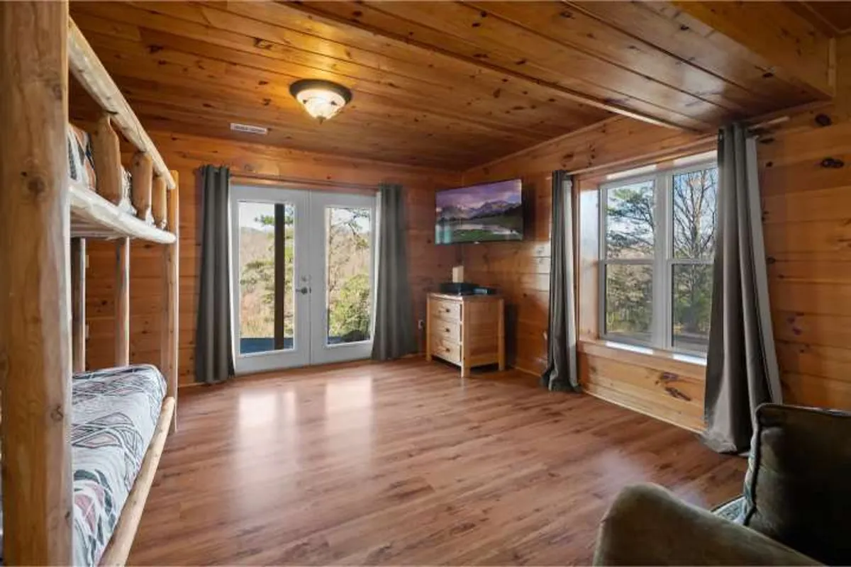 A cozy bunk room features sturdy wooden bunk beds, a television for entertainment, natural wood furniture, and sliding glass doors that open to nature views. The space is family-friendly, with plenty of natural light and a comfortable, inviting atmosphere.