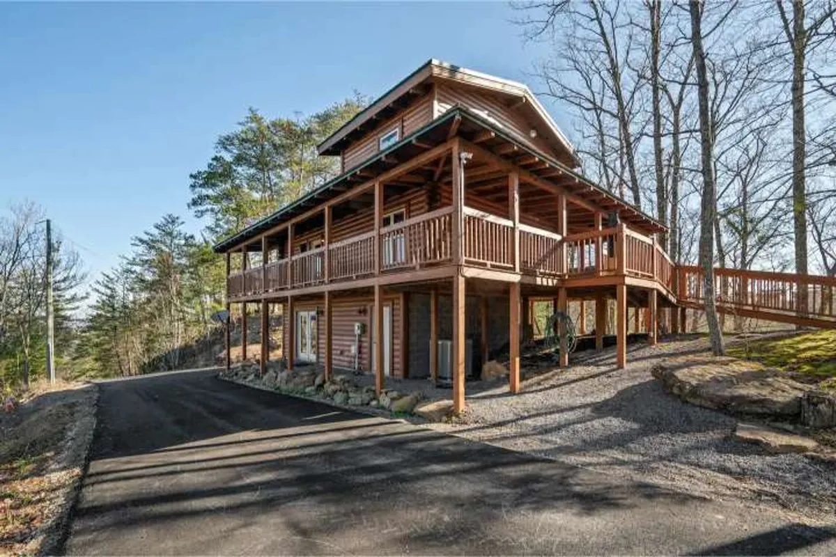 A wooden porch surrounded by trees, featuring large windows and a driveway, offers a tranquil spot for guests to unwind. The space is suitable for gathering around a fire pit or simply relaxing while immersed in nature, with connections to nearby trails for exploration.