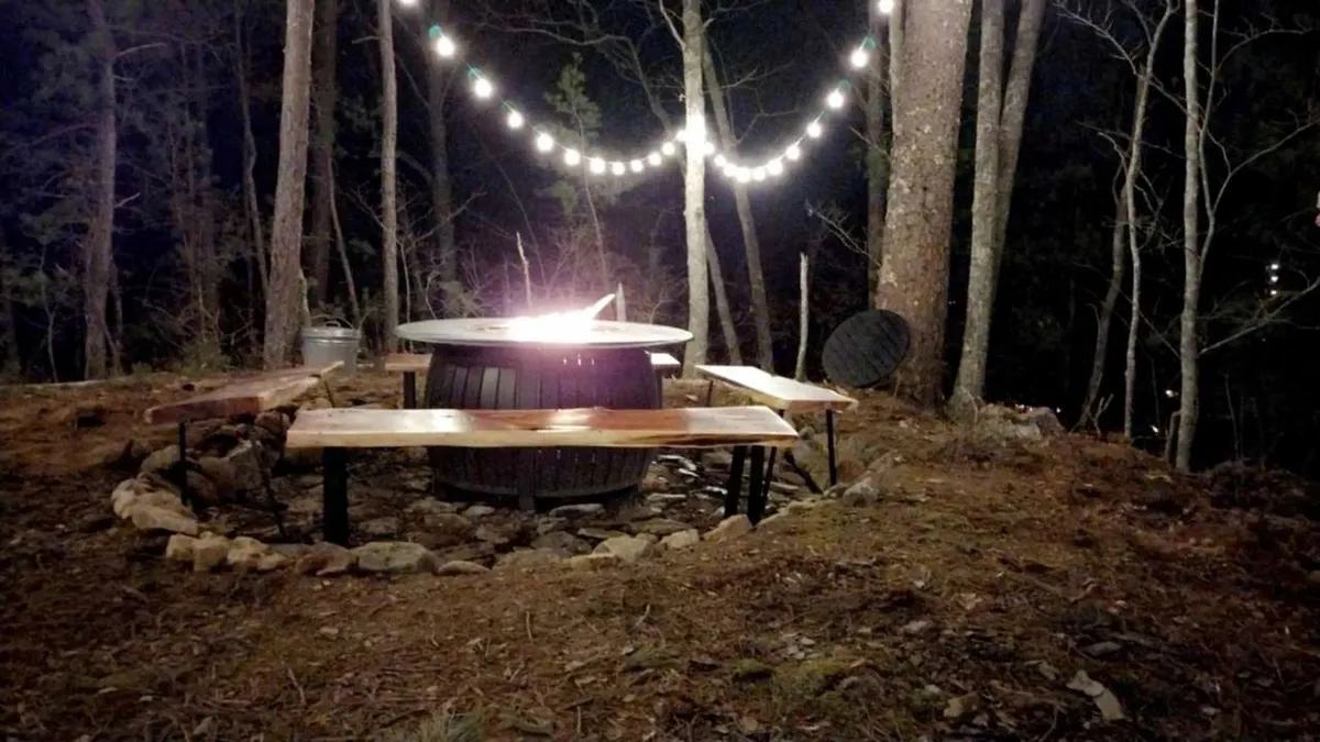 A glowing fire pit surrounded by wooden benches and strung with warm lights, set among trees. The scene radiates warmth and camaraderie, encouraging storytelling, marshmallow toasting, and socializing under the night sky.