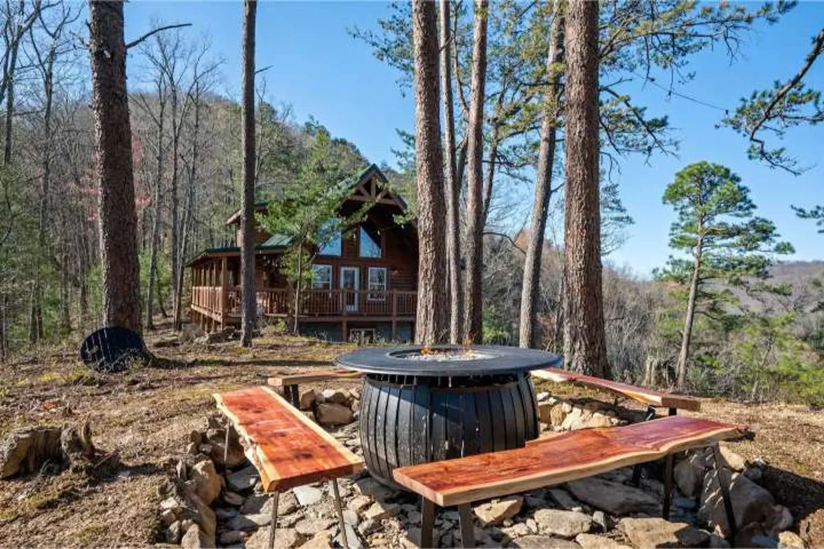 This image captures the exterior of a log cabin surrounded by trees, with a prominent fire pit area and wooden benches for group gatherings. The setting is inviting, with a relaxed feel that hints at evenings spent sharing stories around the fire and enjoying the outdoors together.