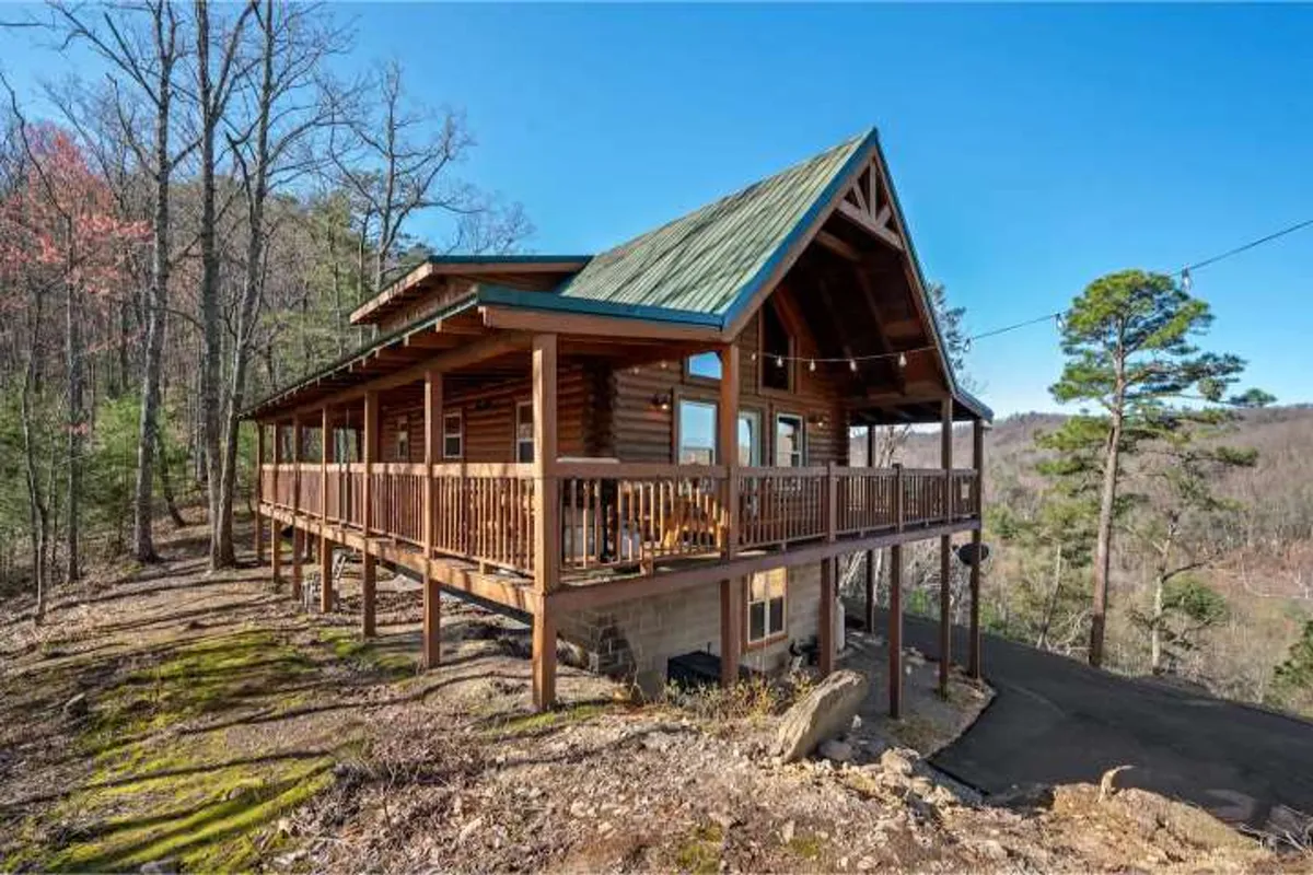 This image shows a cozy log cabin with a wrap-around porch nestled among trees, with a clear view of the mountains in the background. The porch is inviting, perfectly suited for relaxing and soaking in the natural beauty that surrounds the cabin.